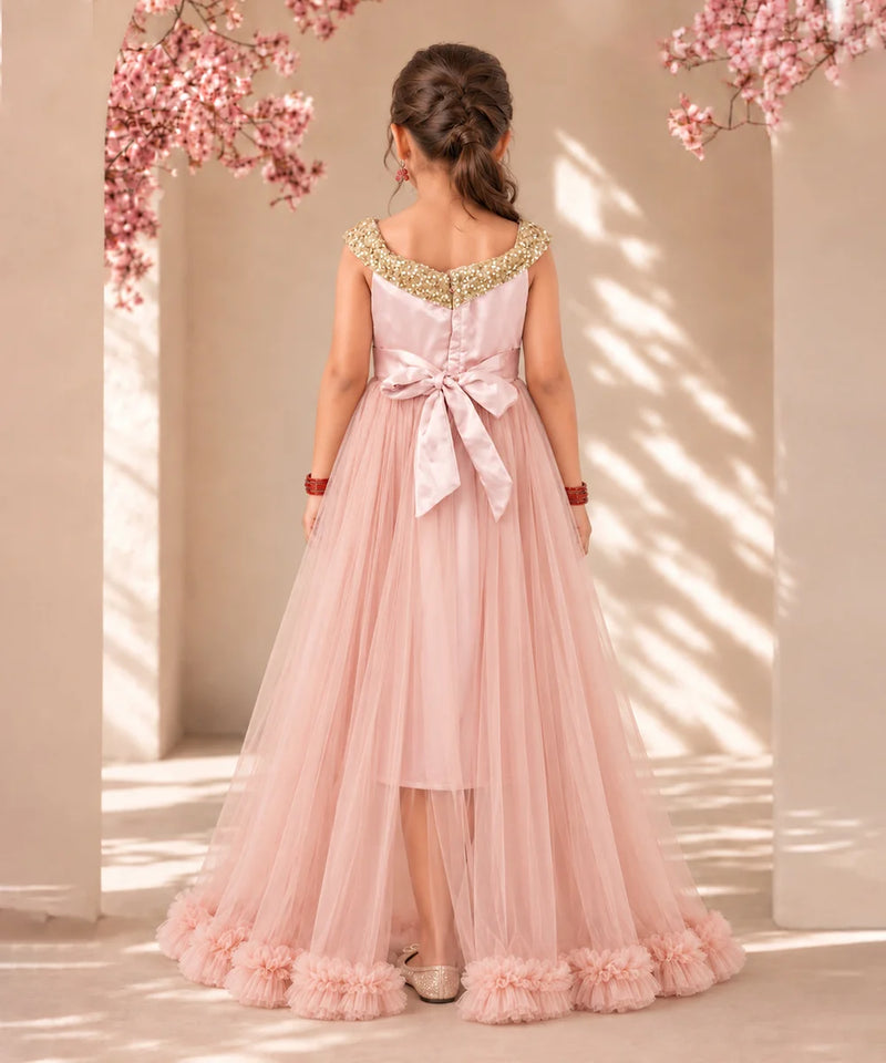 A young girl viewed from behind wearing a formal dusty pink tulle dress with gold beaded shoulder straps, a large satin bow at the back waist, and ruffled trim along the hem, standing in a sunlit architectural setting with pink blossoms.