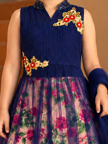 A sleeveless navy blue ethnic dress with a pleated bodice, floral printed tulle skirt, and gold and red embroidered floral details on the chest and waist. The dress is shown on a model against a warm brown background.