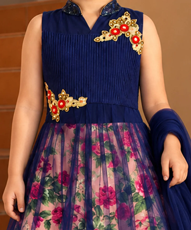 A sleeveless navy blue ethnic dress with a pleated bodice, floral printed tulle skirt, and gold and red embroidered floral details on the chest and waist. The dress is shown on a model against a warm brown background.
