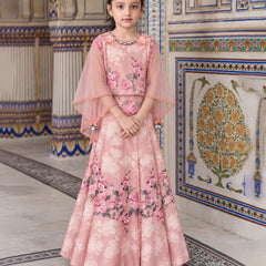 A young girl stands in an ornate indoor setting wearing a pink floral traditional outfit with a sheer draped cape and a long flared skirt. The background features decorative tiled walls and columns.