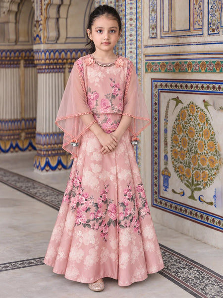 A young girl stands in an ornate indoor setting wearing a pink floral traditional outfit with a sheer draped cape and a long flared skirt. The background features decorative tiled walls and columns.