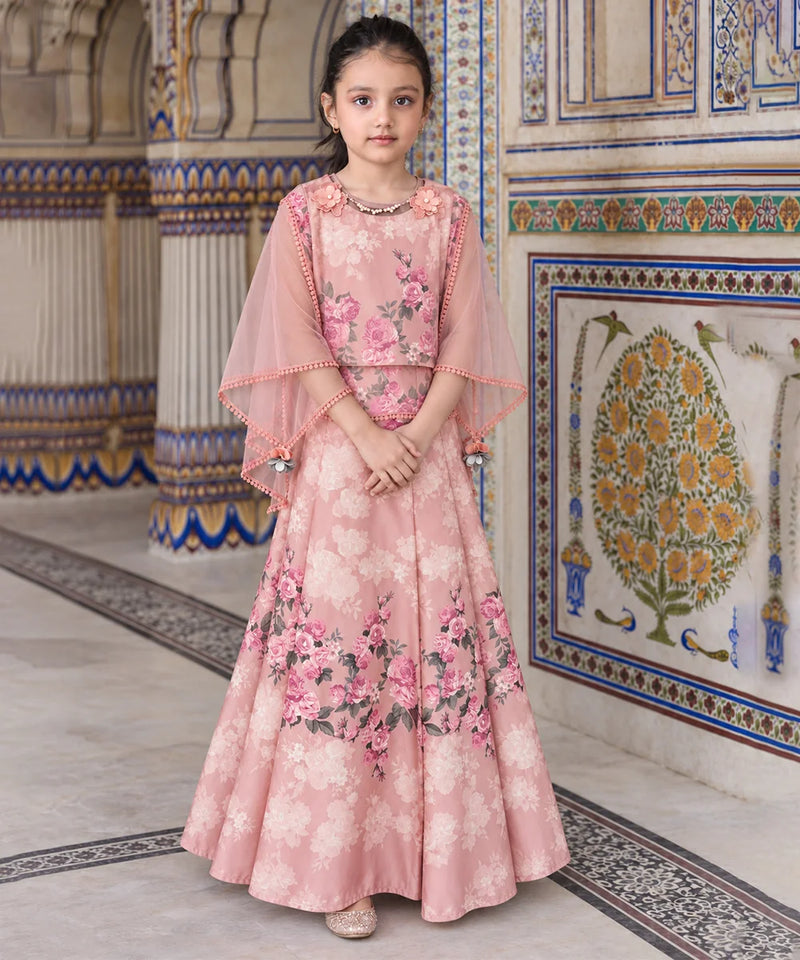 A young girl stands in an ornate indoor setting wearing a pink floral traditional outfit with a sheer draped cape and a long flared skirt. The background features decorative tiled walls and columns.