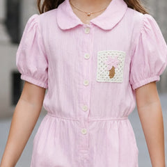 A child wearing a light pink short-sleeve collared dress with puff sleeves, a button front, and a decorative patch pocket on the chest.