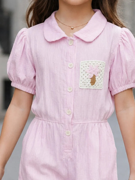 A child wearing a light pink short-sleeve collared dress with puff sleeves, a button front, and a decorative patch pocket on the chest.