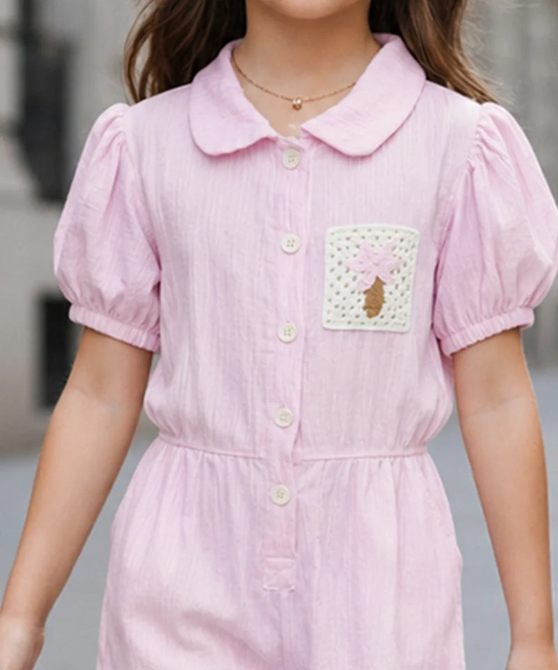 A child wearing a light pink short-sleeve collared dress with puff sleeves, a button front, and a decorative patch pocket on the chest.