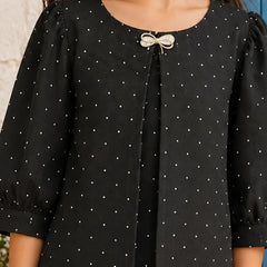 A close-up of a black short-sleeve blouse with a white polka dot pattern, featuring a decorative bow detail at the neckline, worn by a model.