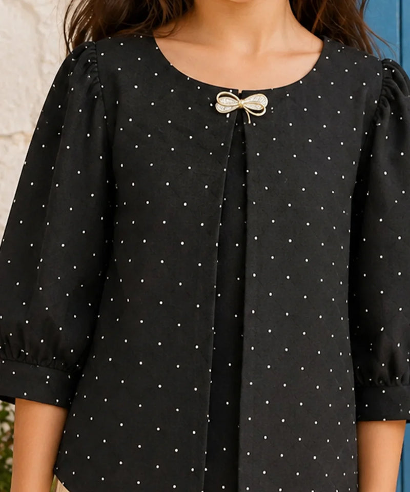 A close-up of a black short-sleeve blouse with a white polka dot pattern, featuring a decorative bow detail at the neckline, worn by a model.