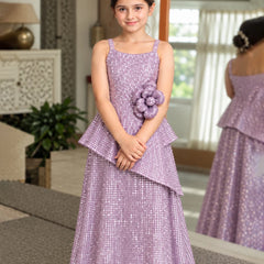 A young girl wearing a lavender sequined formal dress with a peplum waist and a matching flower accessory on the hip, standing in a decorated indoor room with a mirror showing her back.