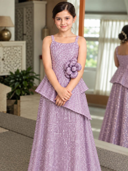 A young girl wearing a lavender sequined formal dress with a peplum waist and a matching flower accessory on the hip, standing in a decorated indoor room with a mirror showing her back.