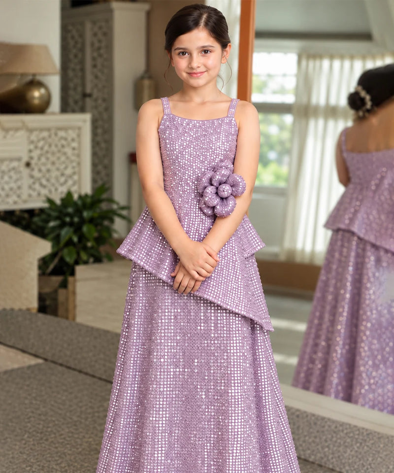 A young girl wearing a lavender sequined formal dress with a peplum waist and a matching flower accessory on the hip, standing in a decorated indoor room with a mirror showing her back.
