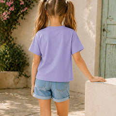 A young girl with blonde pigtails is shown from behind, wearing a lavender purple short-sleeve t-shirt and light blue denim shorts, standing outdoors next to a light-colored stone wall.