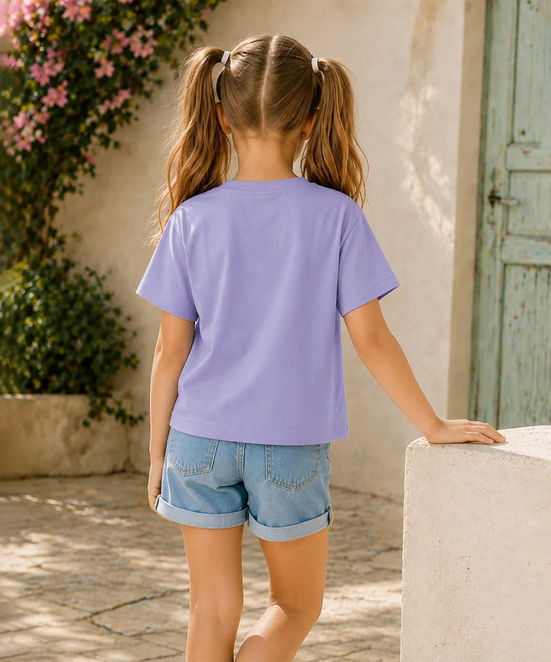 A young girl with blonde pigtails is shown from behind, wearing a lavender purple short-sleeve t-shirt and light blue denim shorts, standing outdoors next to a light-colored stone wall.