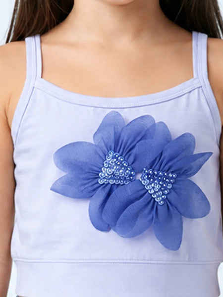 A close-up of a child wearing a light lavender camisole top with a large blue flower applique on the front, featuring blue and silver rhinestone centers.