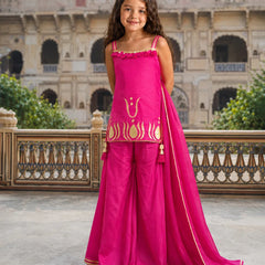 A young girl stands outdoors in front of a historic stone building, wearing a bright pink traditional outfit with gold embroidery and a matching dupatta.