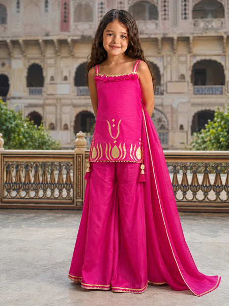 A young girl stands outdoors in front of a historic stone building, wearing a bright pink traditional outfit with gold embroidery and a matching dupatta.