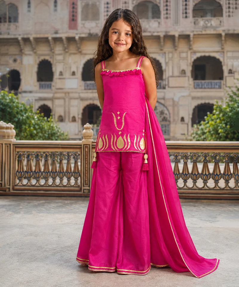 A young girl stands outdoors in front of a historic stone building, wearing a bright pink traditional outfit with gold embroidery and a matching dupatta.