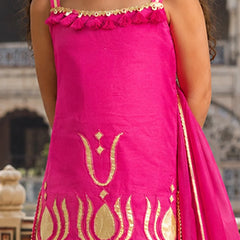 A bright pink traditional outfit with gold embroidery and a matching pink dupatta, shown on a model in front of a blurred architectural background.