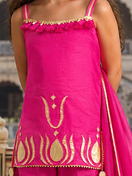A bright pink traditional outfit with gold embroidery and a matching pink dupatta, shown on a model in front of a blurred architectural background.