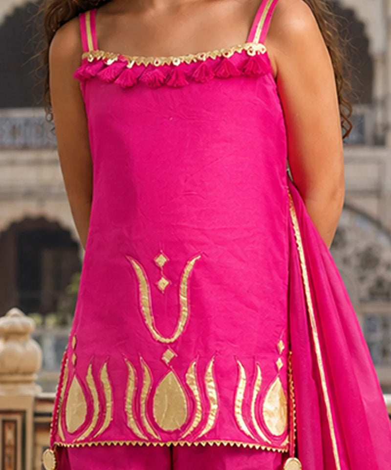 A bright pink traditional outfit with gold embroidery and a matching pink dupatta, shown on a model in front of a blurred architectural background.