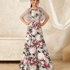 A young girl modeling a long floral printed gown with sheer flutter sleeves, standing in front of a cream-colored textured wall with draped white fabric.