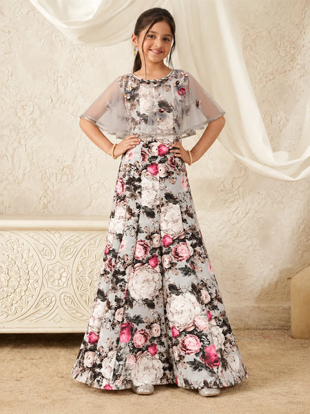 A young girl modeling a long floral printed gown with sheer flutter sleeves, standing in front of a cream-colored textured wall with draped white fabric.