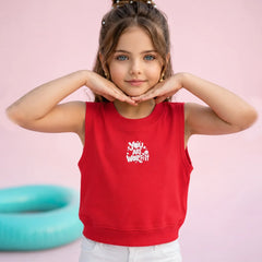 A young girl with her hair in a bun poses in a bright red sleeveless crop top with a white graphic print that reads 'You Are Worth It'. She is wearing white pants against a solid pink background with a teal pool float visible on the left.