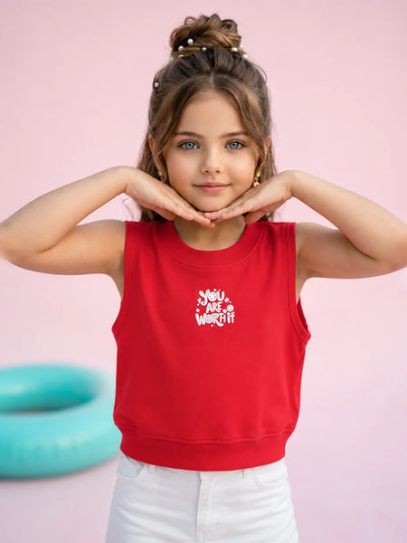 A young girl with her hair in a bun poses in a bright red sleeveless crop top with a white graphic print that reads 'You Are Worth It'. She is wearing white pants against a solid pink background with a teal pool float visible on the left.
