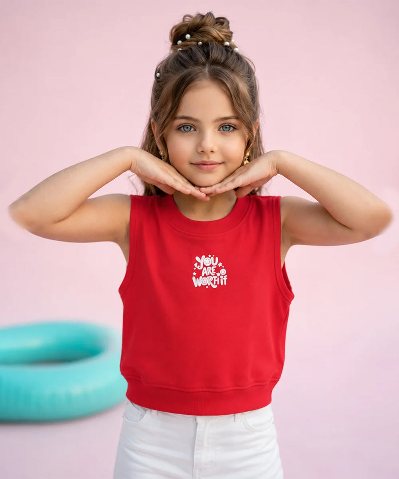 A young girl with her hair in a bun poses in a bright red sleeveless crop top with a white graphic print that reads 'You Are Worth It'. She is wearing white pants against a solid pink background with a teal pool float visible on the left.