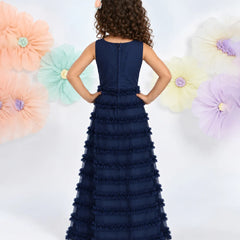 A young girl with curly brown hair is shown from the back wearing a sleeveless navy blue formal gown with a ruffled tulle skirt. She is standing against a white background decorated with large pastel paper flowers.