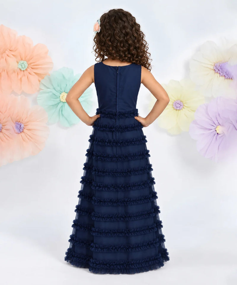 A young girl with curly brown hair is shown from the back wearing a sleeveless navy blue formal gown with a ruffled tulle skirt. She is standing against a white background decorated with large pastel paper flowers.