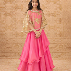 A young girl modeling a bright pink traditional Indian outfit with a layered ruffled skirt, a matching embroidered blouse, and sheer gold-patterned cape sleeves, posed against a patterned beige backdrop.
