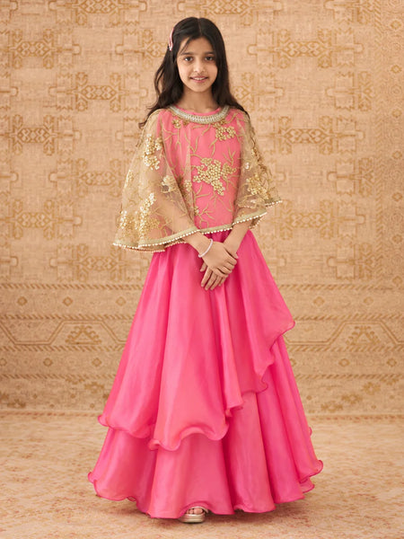 A young girl modeling a bright pink traditional Indian outfit with a layered ruffled skirt, a matching embroidered blouse, and sheer gold-patterned cape sleeves, posed against a patterned beige backdrop.