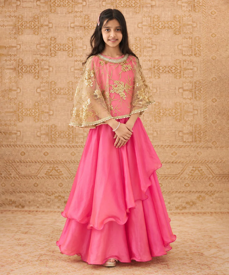 A young girl modeling a bright pink traditional Indian outfit with a layered ruffled skirt, a matching embroidered blouse, and sheer gold-patterned cape sleeves, posed against a patterned beige backdrop.