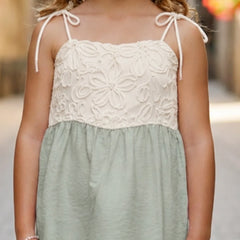 A young girl wearing a sleeveless two-tone top with a cream floral lace bodice and thin shoulder ties, paired with a sage green gathered skirt, shown in an outdoor setting.
