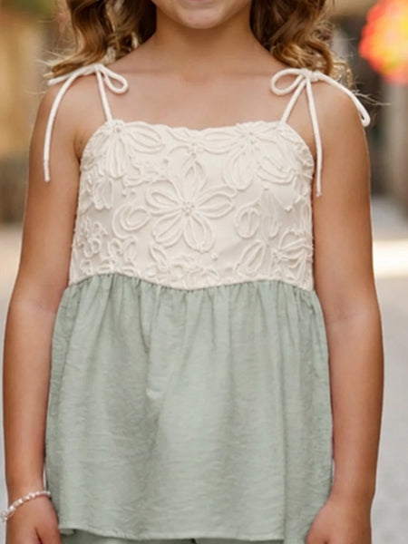 A young girl wearing a sleeveless two-tone top with a cream floral lace bodice and thin shoulder ties, paired with a sage green gathered skirt, shown in an outdoor setting.