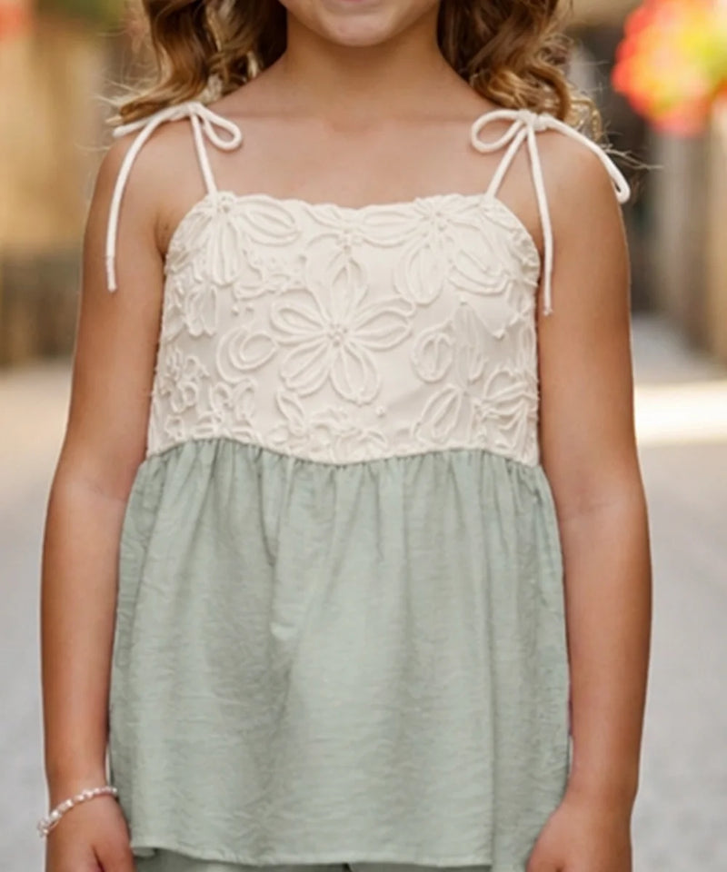 A young girl wearing a sleeveless two-tone top with a cream floral lace bodice and thin shoulder ties, paired with a sage green gathered skirt, shown in an outdoor setting.