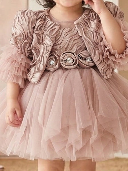 A young child wearing a dusty pink formal dress with a ruffled jacket-style bodice, tulle skirt, and decorative floral rosettes at the waist.
