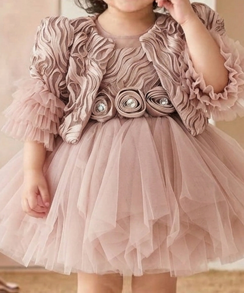 A young child wearing a dusty pink formal dress with a ruffled jacket-style bodice, tulle skirt, and decorative floral rosettes at the waist.