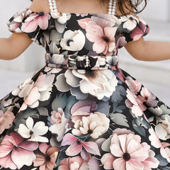 A close-up of a child wearing an off-the-shoulder floral dress with a matching belt and pearl-like shoulder accents. The dress has a black background with large pink, white, and gray flowers, and the child is also wearing pearl necklaces.