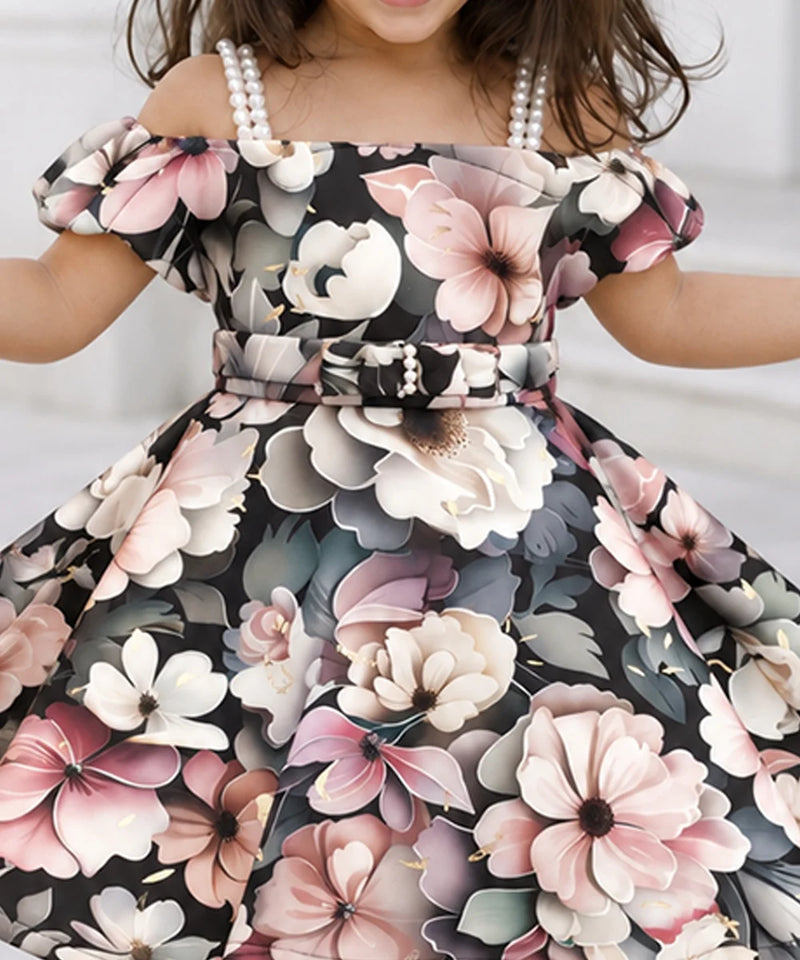 A close-up of a child wearing an off-the-shoulder floral dress with a matching belt and pearl-like shoulder accents. The dress has a black background with large pink, white, and gray flowers, and the child is also wearing pearl necklaces.