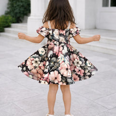 A young girl viewed from behind wearing a black off-the-shoulder floral dress with pink, white, and gray flowers, standing on a light stone patio in front of a white building.
