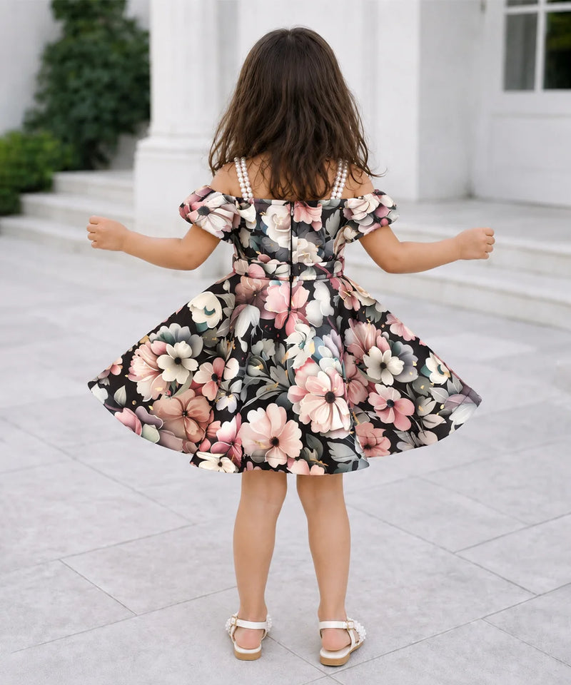 A young girl viewed from behind wearing a black off-the-shoulder floral dress with pink, white, and gray flowers, standing on a light stone patio in front of a white building.