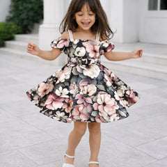 A young girl with long brown hair is wearing a floral off-the-shoulder dress with a flared skirt and white strappy heels, standing outdoors in front of a white building with columns.
