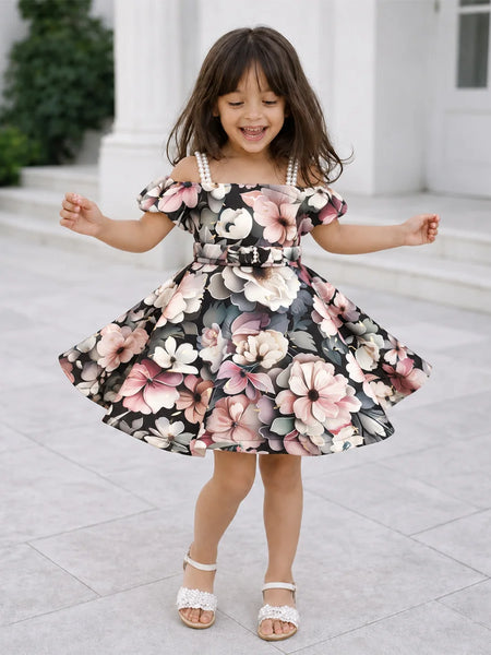 A young girl with long brown hair is wearing a floral off-the-shoulder dress with a flared skirt and white strappy heels, standing outdoors in front of a white building with columns.
