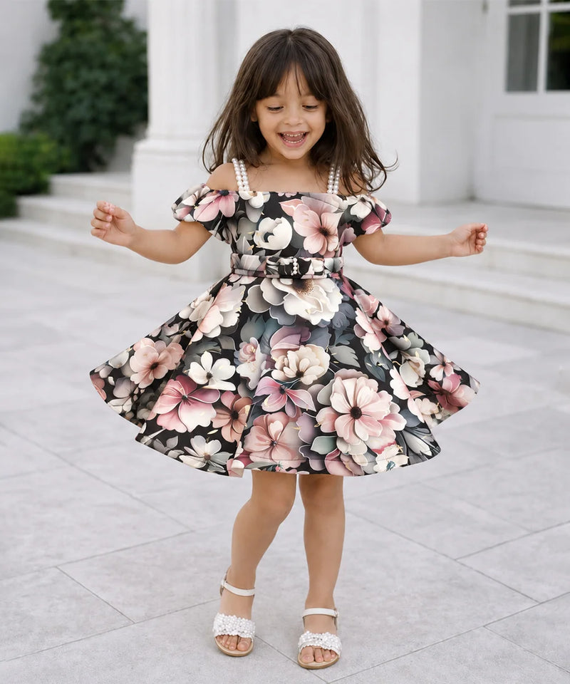 A young girl with long brown hair is wearing a floral off-the-shoulder dress with a flared skirt and white strappy heels, standing outdoors in front of a white building with columns.