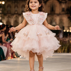 A young girl smiling and walking down a fashion runway wearing a light pink high-low party dress with floral embroidery and tulle details, with a blurred indoor venue in the background.