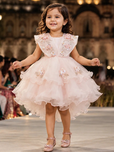 A young girl smiling and walking down a fashion runway wearing a light pink high-low party dress with floral embroidery and tulle details, with a blurred indoor venue in the background.