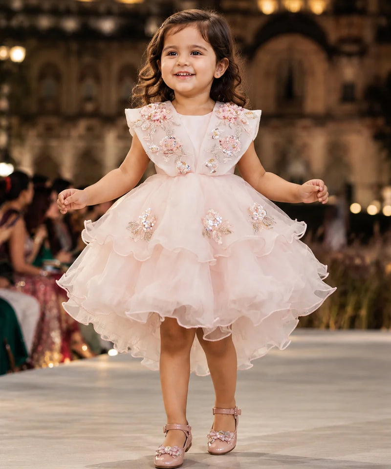 A young girl smiling and walking down a fashion runway wearing a light pink high-low party dress with floral embroidery and tulle details, with a blurred indoor venue in the background.