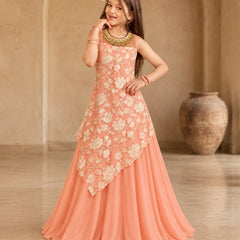 A young girl modeling a peach-colored sleeveless ethnic gown with cream floral embroidery and a draped asymmetrical overlay, set against a warm beige architectural background with a large clay pot.
