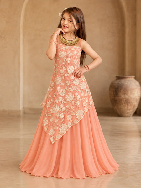 A young girl modeling a peach-colored sleeveless ethnic gown with cream floral embroidery and a draped asymmetrical overlay, set against a warm beige architectural background with a large clay pot.